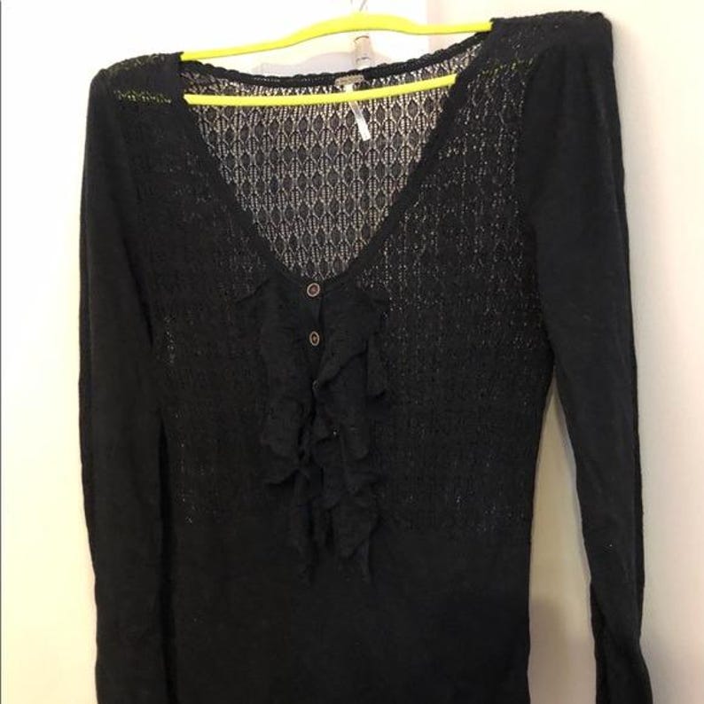 Pu&ograve; includere: Maglione nero a maniche lunghe con scollo a V e un delicato motivo a pizzo sulla parte superiore del petto. Il maglione presenta un dettaglio a balze sul davanti ed &egrave; allacciato con due bottoni. L'indumento &egrave; appeso a una gruccia gialla.