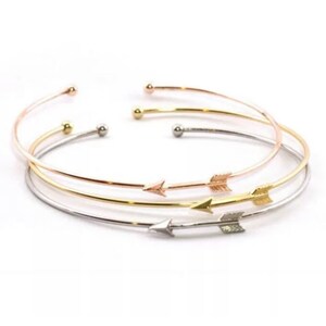 May include: Three delicate gold, silver, and rose gold bangle bracelets with arrow charms. The bracelets are stacked on top of each other.