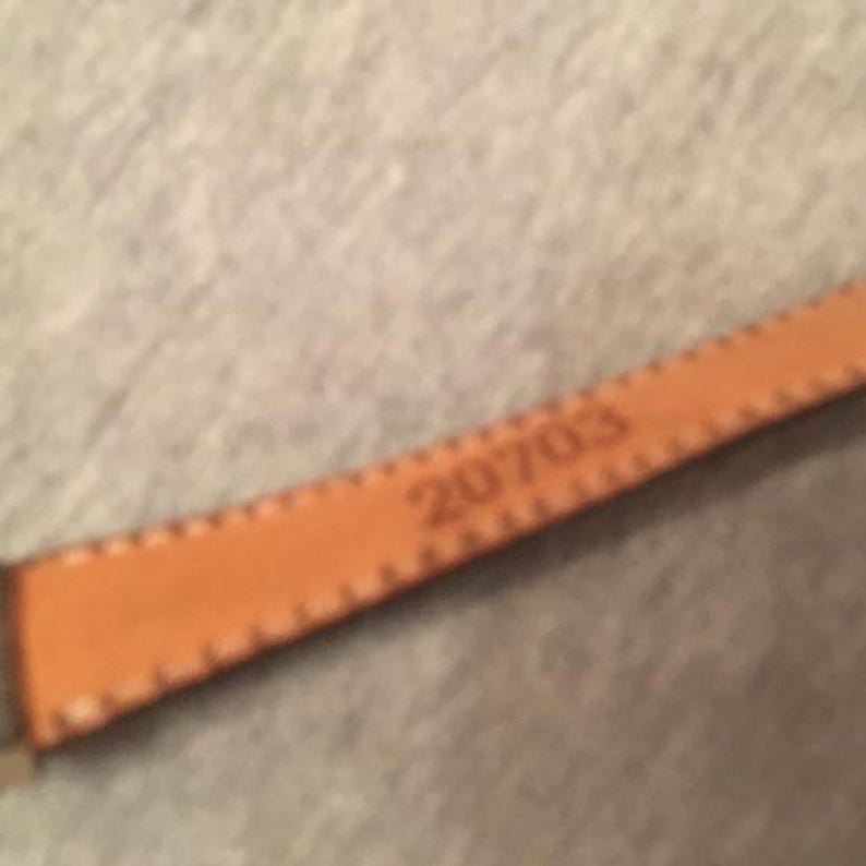 Op de afbeelding: Close-up van een bruinleren bandje met het nummer "20703" erin ge&euml;mbosseerd. De band heeft een gestikte rand en lijkt deel uit te maken van een groter item, mogelijk een horlogebandje of riem.