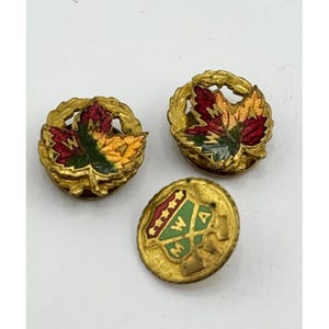 Puede incluir: Tres broches vintage de metal dorado. Dos broches presentan un diseño de hoja de arce con esmalte rojo, verde y amarillo. El tercer broche es redondo con las letras "MWA" y un diseño verde y rojo.