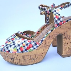 Puede incluir: Sandalias de plataforma con tacón de cuña de corcho. Las sandalias de punta abierta presentan un estampado de cuadros vichy en blanco y negro con detalles de fresas y flores. La correa del tobillo tiene un detalle de lazo. El tacón mide unos 10 cm.