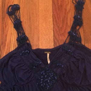 Puede incluir: Una blusa azul oscuro con intrincadas correas tejidas y un escote en V. La blusa presenta adornos decorativos alrededor del escote y en las correas. La tela parece suave y fluida, con un brillo sutil. La blusa es sin mangas.