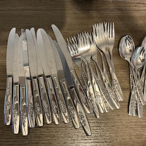 Pode incluir: Uma coleção de talheres de aço inoxidável, incluindo facas, garfos e colheres, dispostos sobre uma superfície de madeira. Os talheres apresentam um design floral nas alças, adicionando um toque decorativo ao conjunto. O conjunto inclui facas, garfos e colheres.