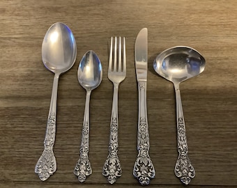 Può includere: Un set di cinque posate color argento: un cucchiaio da zuppa, un cucchiaino, una forchetta, un coltello e un mestolo. Ogni pezzo presenta un motivo floreale decorativo sul manico. Le posate sono disposte su una superficie di legno.