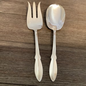 Puede incluir: Un juego de tenedor y cuchara de servir plateados. El tenedor tiene tres púas y la cuchara tiene un cuenco grande y ovalado. Ambos utensilios tienen mangos decorativos y alargados con un diseño en forma de hoja.