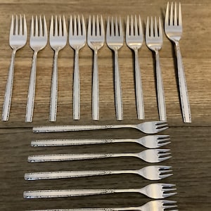 Könnte beinhalten: Eine Sammlung silberner Gabeln, die auf einer Holzoberfläche angeordnet sind. Die Gabeln haben ein dekoratives Muster auf den Griffen. Das Bild zeigt verschiedene Größen und Stile von Gabeln, passend für eine formelle Tischdekoration. Die Gabeln sind aus glänzendem Metall.