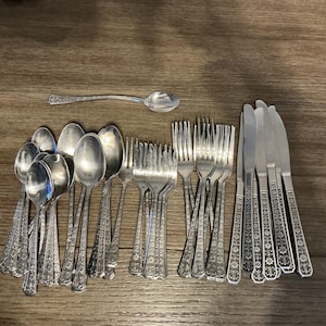 Op de afbeelding: Een verzameling zilverkleurig bestek, waaronder lepels, vorken en messen, gerangschikt op een houten oppervlak. Het bestek heeft sierlijke bloemendetails op de handvatten. De set bevat verschillende maten lepels, vorken en messen, geschikt voor een formele setting.