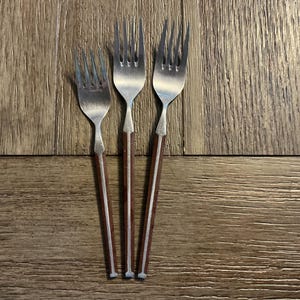 May include: Three silver forks with brown wooden handles are arranged on a wooden surface. The forks have a classic design, with tines and a handle that tapers to a point. The wooden handles add a touch of warmth to the silverware.