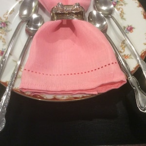 May include: Four silver spoons are wrapped in a pink linen napkin with a silver napkin ring. The spoons are on a white plate with a floral pattern.