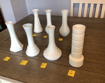 May include: A collection of seven white glass vases with various designs. The vases are arranged on a wooden table, showcasing different shapes and textures, including ribbed, textured, and geometric patterns. The vases vary in height and width.