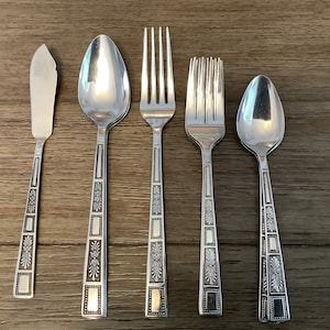 Puede incluir: Cinco piezas de cubiertos, incluyendo un cuchillo para mantequilla, dos cucharas y dos tenedores, todos con un acabado plateado y un diseño ornamentado.