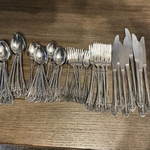 Op de afbeelding: Een verzameling sierlijk zilverkleurig bestek, waaronder lepels, vorken en messen, gerangschikt op een houten oppervlak. Het zilverwerk heeft gedetailleerde decoratieve handvatten. De opstelling suggereert een formele eetsituatie.