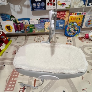May include: A white, plush, oval-shaped baby bouncer with a gray base and a strap. The bouncer is on a patterned play mat with a toy house and other toys.