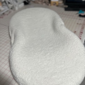 May include: A white, fluffy, oval-shaped cushion or pillow. The cushion is made of a soft, plush material and has a slightly textured surface.