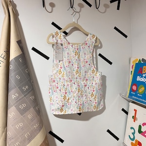 Puede incluir: Un delantal blanco sin mangas con estampado floral con flores rosas, amarillas y verdes. El delantal tiene un pequeño bolsillo en la parte delantera y está colgado de una percha de madera.