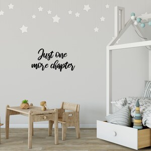 K&ouml;nnte beinhalten: Ein wei&szlig;es Holzbettgestell mit einem hausf&ouml;rmigen Kopfteil und einer wei&szlig;en Schublade darunter. Das Bett ist mit einer wei&szlig;-grauen gestreiften Decke, einem grauen Kissen mit einem Pandagesicht und einem bunten Holz-Stapelspielzeug dekoriert. Die Wand hinter dem Bett hat einen schwarzen Vinyl-Aufkleber mit der Aufschrift "Just one more chapter".