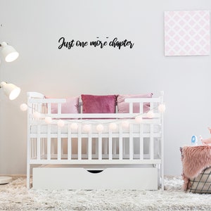 K&ouml;nnte beinhalten: Ein wei&szlig;es Babybett mit einer wei&szlig;en Matratze und einer wei&szlig;en Schublade darunter. Das Bett ist mit einer Lichterkette aus wei&szlig;en Lichtern und rosa Kissen dekoriert. Die Wand hinter dem Babybett hat ein schwarzes Schild mit der Aufschrift "Just one more chapter".