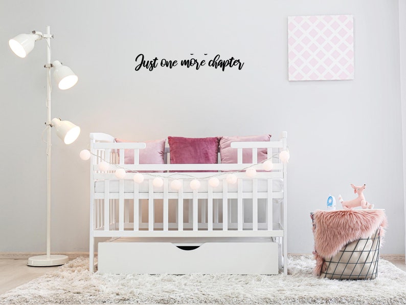 K&ouml;nnte beinhalten: Ein wei&szlig;es Babybett mit einer wei&szlig;en Matratze und einer wei&szlig;en Schublade darunter. Das Bett ist mit einer Lichterkette aus wei&szlig;en Lichtern und rosa Kissen dekoriert. Die Wand hinter dem Babybett hat ein schwarzes Schild mit der Aufschrift "Just one more chapter".