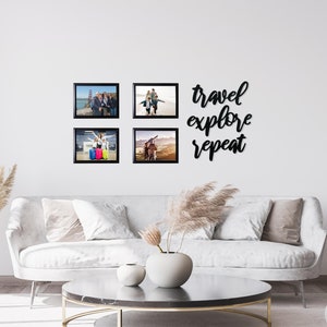 May include: Four black framed photos of people on a white wall with the text "travel explore repeat" in black script above the photos. The photos are of people traveling and enjoying their vacations. The photos are arranged in a square shape. There is a white couch and a coffee table in front of the wall.