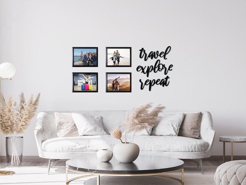 May include: Four black framed photos of people on a white wall with the text "travel explore repeat" in black script above the photos. The photos are of people traveling and enjoying their vacations. The photos are arranged in a square shape. There is a white couch and a coffee table in front of the wall.