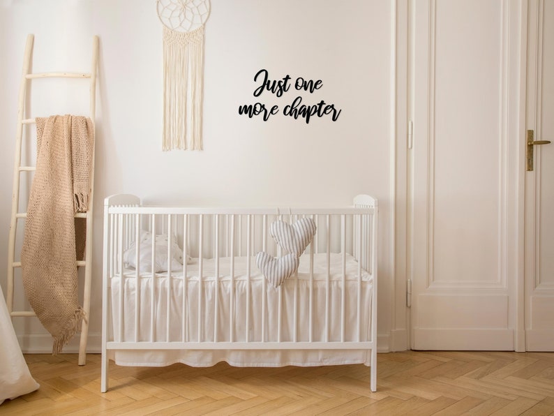 K&ouml;nnte beinhalten: Wei&szlig;es Babybett mit einem wei&szlig;en R&uuml;schenrock und einem schwarzen Wandaufkleber mit der Aufschrift "Just one more chapter".