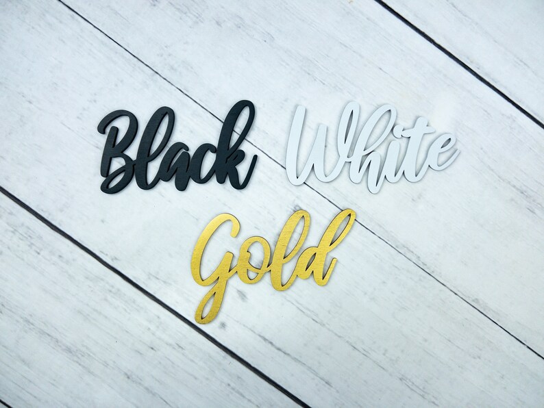 May include: Three wooden words, "Black", "White", and "Gold", are displayed on a white woodgrain background. The words are cut out of wood and painted in their respective colors.