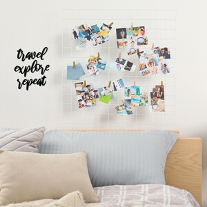 May include: A white grid wall decor with a black metal sign that reads "travel explore repeat" and a collection of photos hanging on it with clothespins. The wall decor is above a bed with a light blue pillow and a tan pillow.