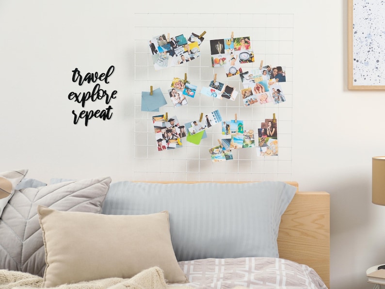 May include: A white grid wall decor with a black metal sign that reads "travel explore repeat" and a collection of photos hanging on it with clothespins. The wall decor is above a bed with a light blue pillow and a tan pillow.