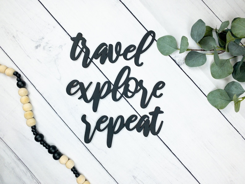 May include: Black wooden sign with the text "travel explore repeat" on a white wood background with a black and white beaded garland and a sprig of eucalyptus.