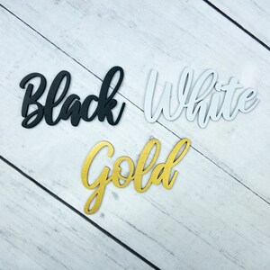 K&ouml;nnte beinhalten: Drei Holzw&ouml;rter, "Black", "White" und "Gold", werden auf einem wei&szlig;en Holzmaserungshintergrund angezeigt. Die W&ouml;rter sind aus Holz ausgeschnitten und in ihren jeweiligen Farben bemalt.