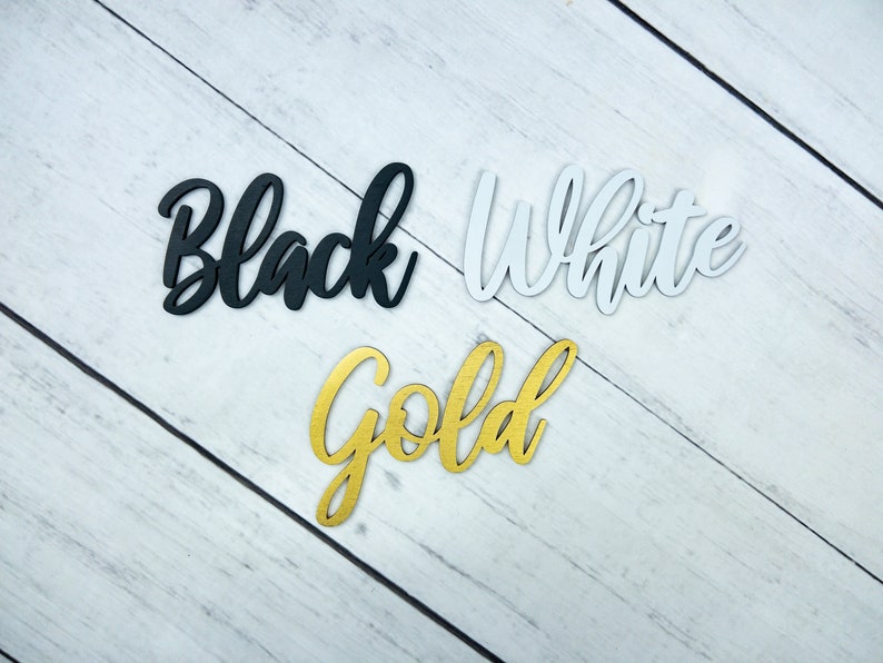 K&ouml;nnte beinhalten: Drei Holzw&ouml;rter, "Black", "White" und "Gold", werden auf einem wei&szlig;en Holzmaserungshintergrund angezeigt. Die W&ouml;rter sind aus Holz ausgeschnitten und in ihren jeweiligen Farben bemalt.