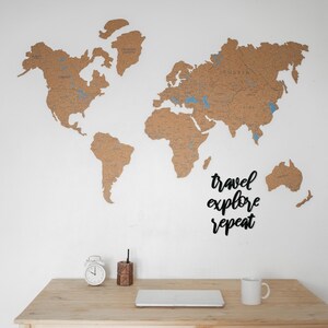 May include: A corkboard world map with blue pins marking locations. The map is mounted on a white wall with a wooden desk below. The desk has a white alarm clock, a wooden pencil holder, a laptop, and a white coffee mug. The text "travel explore repeat" is written in black on the wall.