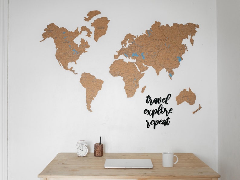 May include: A corkboard world map with blue pins marking locations. The map is mounted on a white wall with a wooden desk below. The desk has a white alarm clock, a wooden pencil holder, a laptop, and a white coffee mug. The text "travel explore repeat" is written in black on the wall.