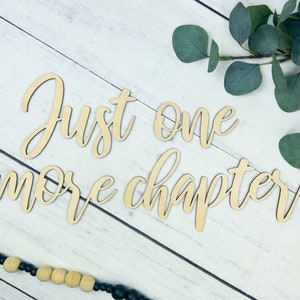 K&ouml;nnte beinhalten: Ein Holzschild mit dem Text "Just one more chapter" in kursivschrift. Das Schild befindet sich auf einem wei&szlig;en Holzgrund mit einem Zweig Eukalyptusbl&auml;tter.