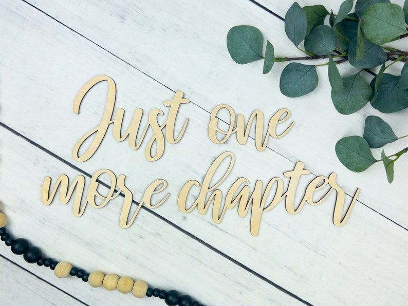 K&ouml;nnte beinhalten: Ein Holzschild mit dem Text "Just one more chapter" in kursivschrift. Das Schild befindet sich auf einem wei&szlig;en Holzgrund mit einem Zweig Eukalyptusbl&auml;tter.