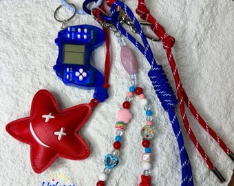 May include: A collection of colorful keychains and accessories. Includes a blue handheld game, a red star-shaped charm with white crosses, and beaded strands with various charms. The keychains have red, blue, and white cords.