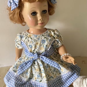 May include: A vintage doll with red hair, blue eyes, and freckles, wearing a floral dress with a blue gingham sash and trim. The doll has blue gingham bows in her hair and a pearl bracelet. The dress has a white lace trim.