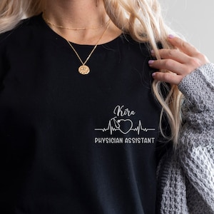 Könnte beinhalten: Ein schwarzes T-Shirt mit einem weißen Grafikdesign, auf dem "Kira Physician Assistant" mit einem Herzen und einer Herzschlaglinie steht.