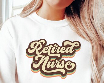 Puede incluir: Una sudadera blanca con el texto "Retired Nurse" en una fuente retro con colores marrón, amarillo y verde.