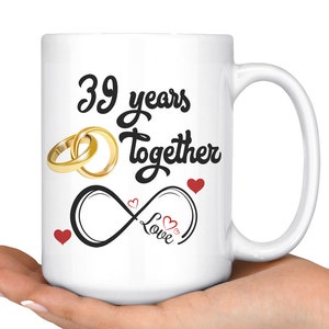 Puede incluir: Taza de cerámica blanca con un diseño negro que dice "39 años juntos" con dos anillos dorados y un símbolo de infinito con la palabra "love" dentro. Hay pequeños corazones rojos alrededor del diseño.