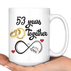 May include: White ceramic coffee mug with a black inscription that reads "53 years together" with a gold ring design and an infinity symbol with the word "love" inside. There are small red hearts around the design.