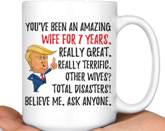 May include: White ceramic coffee mug with a black and red graphic of a man with a red tie and a white shirt. The text on the mug reads "You've been an amazing wife for 7 years. Really great. Really terrific. Other wives? Total disasters! Believe me, ask anyone."