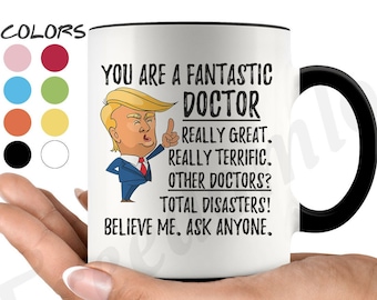 May include: White ceramic coffee mug with a black handle. The mug features a cartoon image of a man with a red tie and a black suit. The text on the mug reads "You are a fantastic doctor. Really great. Really terrific. Other doctors? Total disasters! Believe me. Ask anyone."