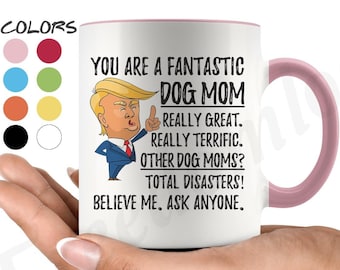 May include: White ceramic coffee mug with a pink rim. The mug features a cartoon image of Donald Trump with the text "You are a fantastic dog mom. Really great. Really terrific. Other dog moms? Total disasters! Believe me. Ask anyone."