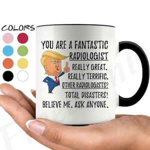 May include: White coffee mug with a black handle. The mug features a cartoon image of Donald Trump with the text "You are a fantastic Radiologist. Really great. Really terrific. Other Radiologists? Total disasters! Believe me. Ask anyone."