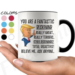 May include: White ceramic coffee mug with a black handle. The mug features a cartoon image of a man with blonde hair and a red tie. The text on the mug reads "You are a fantastic deckhand. Really great. Really terrific. Other deckhands? Total disasters! Believe me. Ask anyone."
