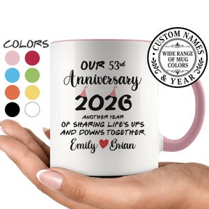 May include: A white ceramic mug with a pink handle and interior, featuring the text "OUR 53rd Anniversary 2026 ANOTHER YEAR OF SHARING LIFE'S UPS AND DOWNS TOGETHER. Emily Brian" with a small red heart and two party hats printed on the front.