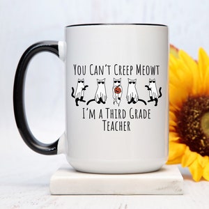 Puede incluir: Taza de cerámica blanca con asa y borde negros, con el texto "You Can't Creep Meowt I'm a Third Grade Teacher". El diseño incluye gatos de dibujos animados con gafas de sol y disfraces de fantasma. Una flor de girasol amarilla en el fondo.