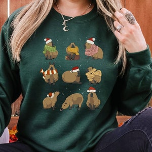 Op de afbeelding: Een donkergroen sweatshirt met een feestelijk ontwerp met meerdere capibara's die kerstmanmutsen en sjaals dragen. De capibara's zijn in een rasterpatroon op de voorkant van het sweatshirt gerangschikt.