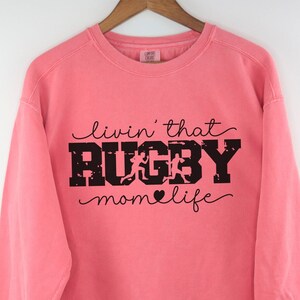 Puede incluir: Una sudadera rosa con el texto "livin' that RUGBY momlife" en negro. El texto está en una fuente desgastada con un pequeño corazón entre "mom" y "life".